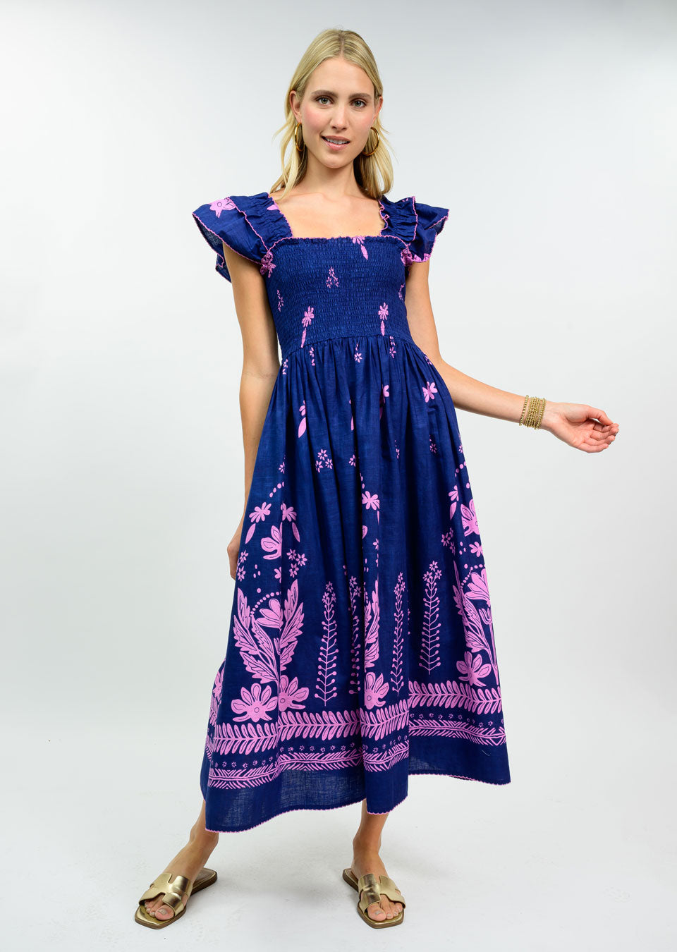 Woman wearing a navy blue 100% cotton midi dress with a smocked bodice, pink floral embroidery, and ruffle cap sleeves