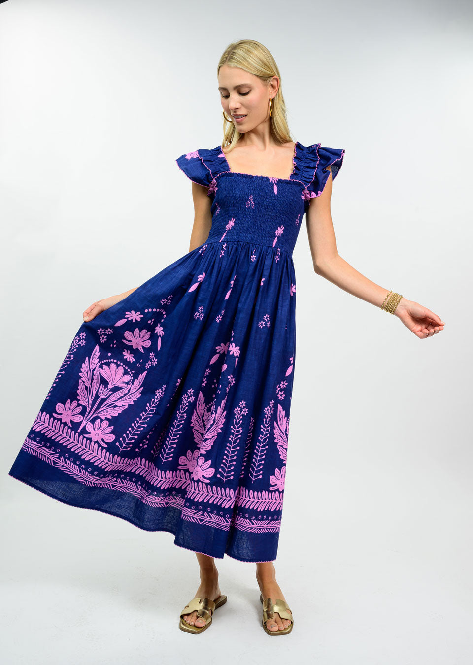 Woman wearing a navy blue 100% cotton midi dress with a smocked bodice, pink floral embroidery, and ruffle cap sleeves.