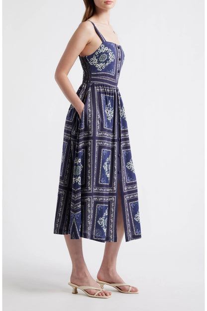 Side view of a woman wearing a navy blue bandana-patterned cotton-linen midi dress with a square neckline, adjustable straps, and a button-front skirt.