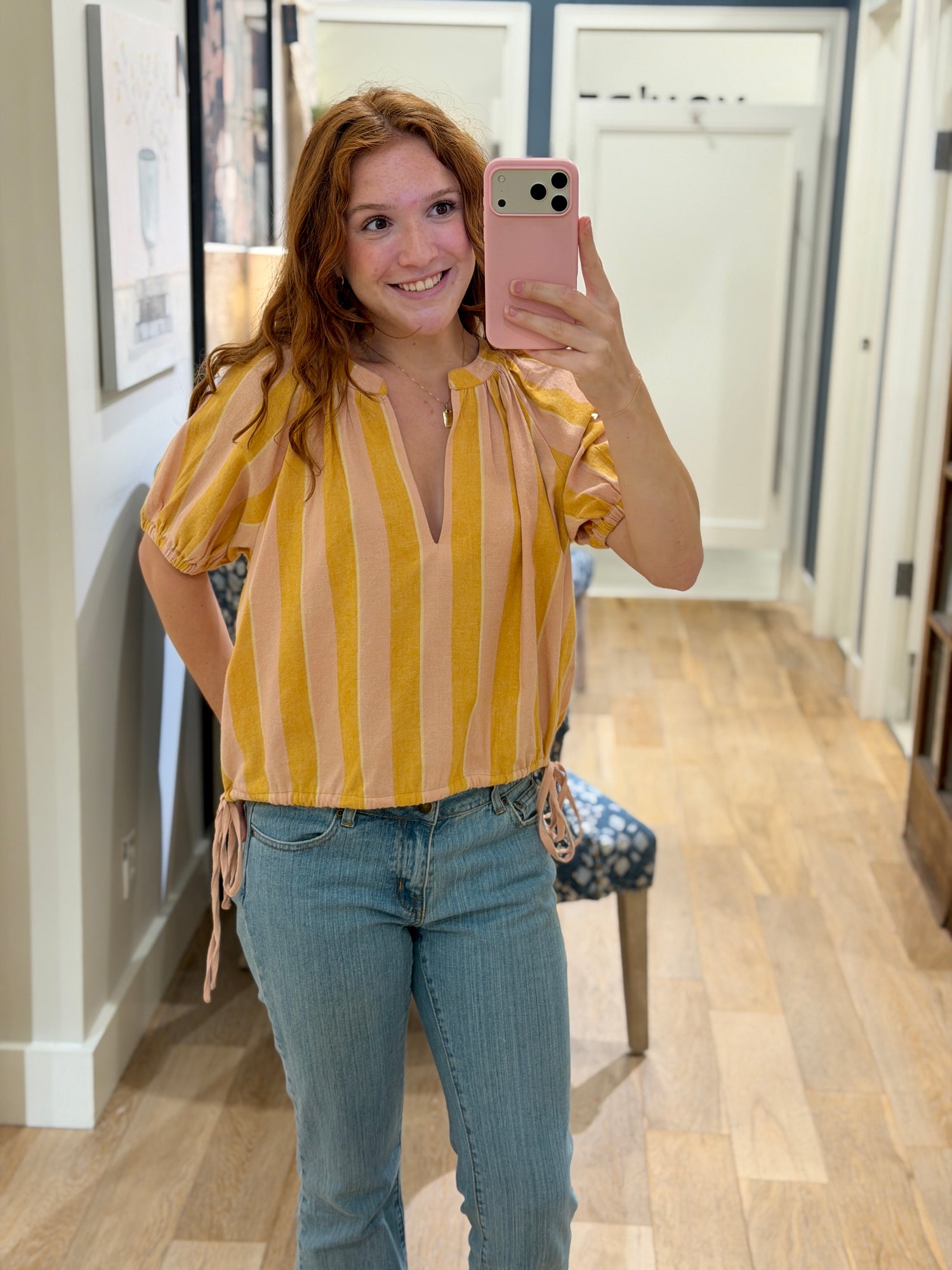 Smiling young woman taking a mirror selfie wearing a peach and mustard yellow striped cotton blouse with blue jeans.