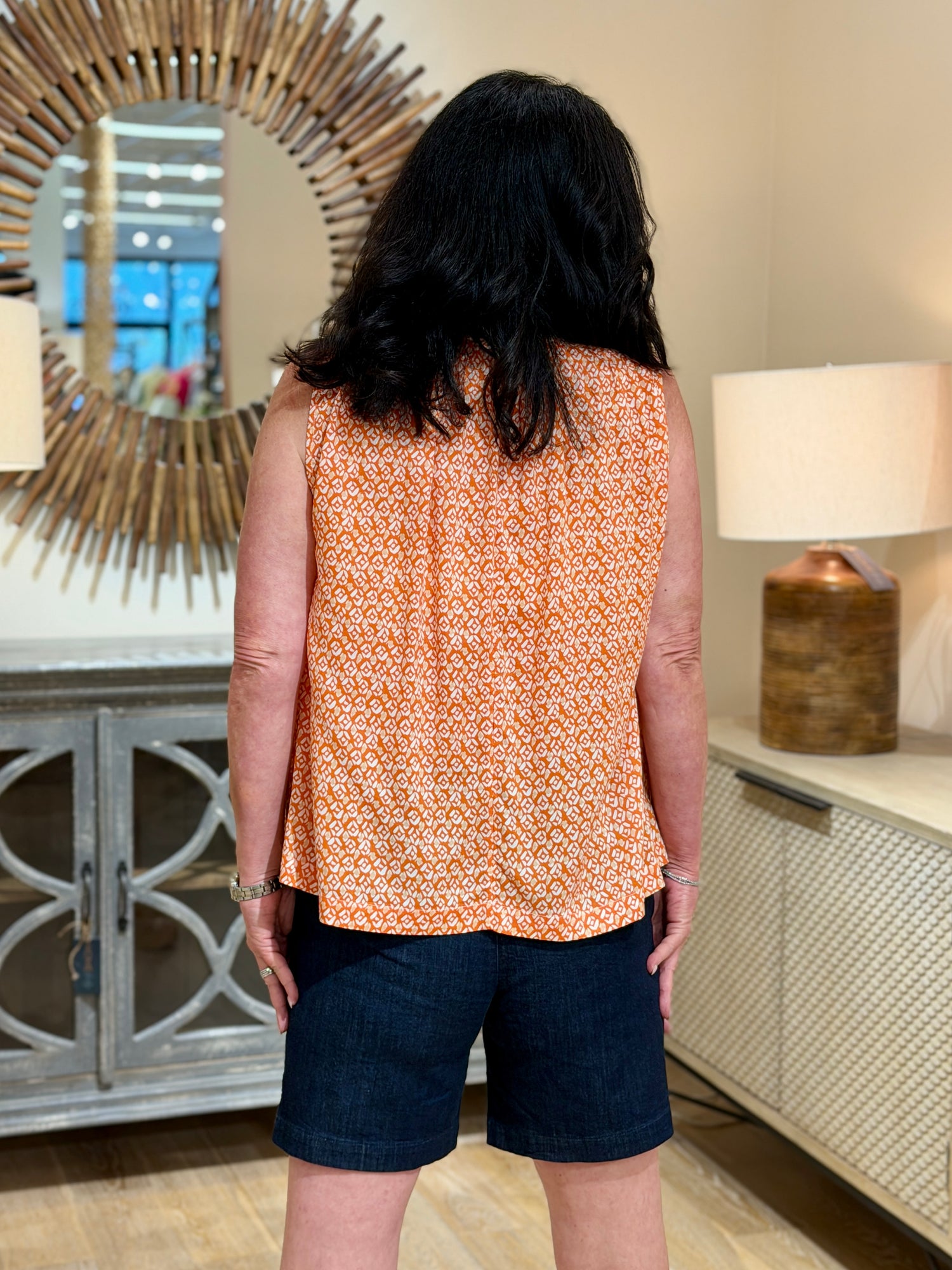 Close up rear view of a woman with dark hair smiling while wearing an orange and white geometric print sleeveless rayon blouse paired with dark denim shorts.