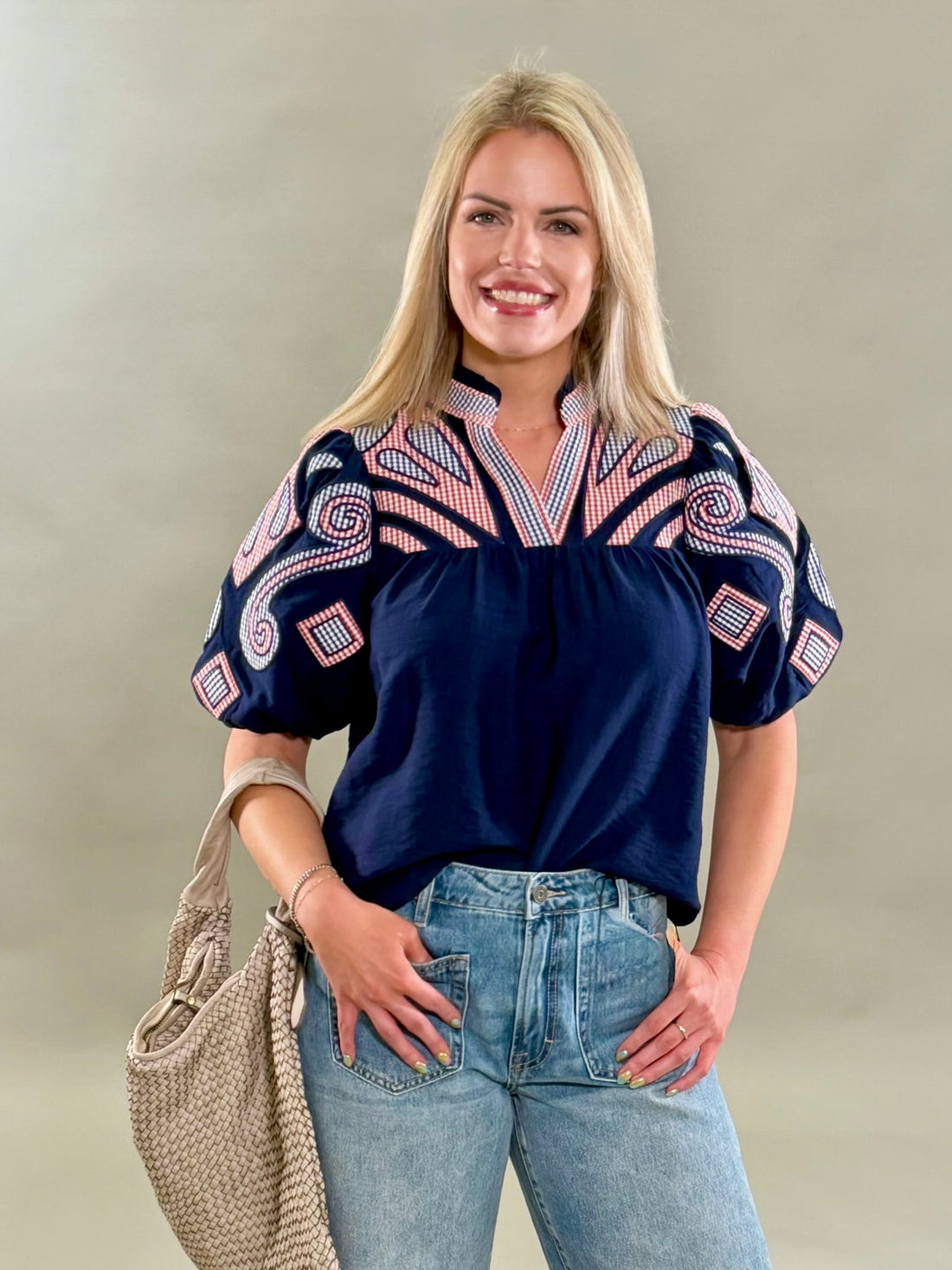 A close up of a smiling blonde woman wearing a textured navy blouse with intricate red, white, and blue plaid embroidery on the yoke and puff sleeves, paired with light-wash jeans.