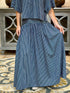 A person wearing a matching blue and white vertically striped two-piece outfit, featuring a short-sleeve cropped top and a high-waisted, full-length skirt standing on a patterned rug in front of a gray wooden cabinet.