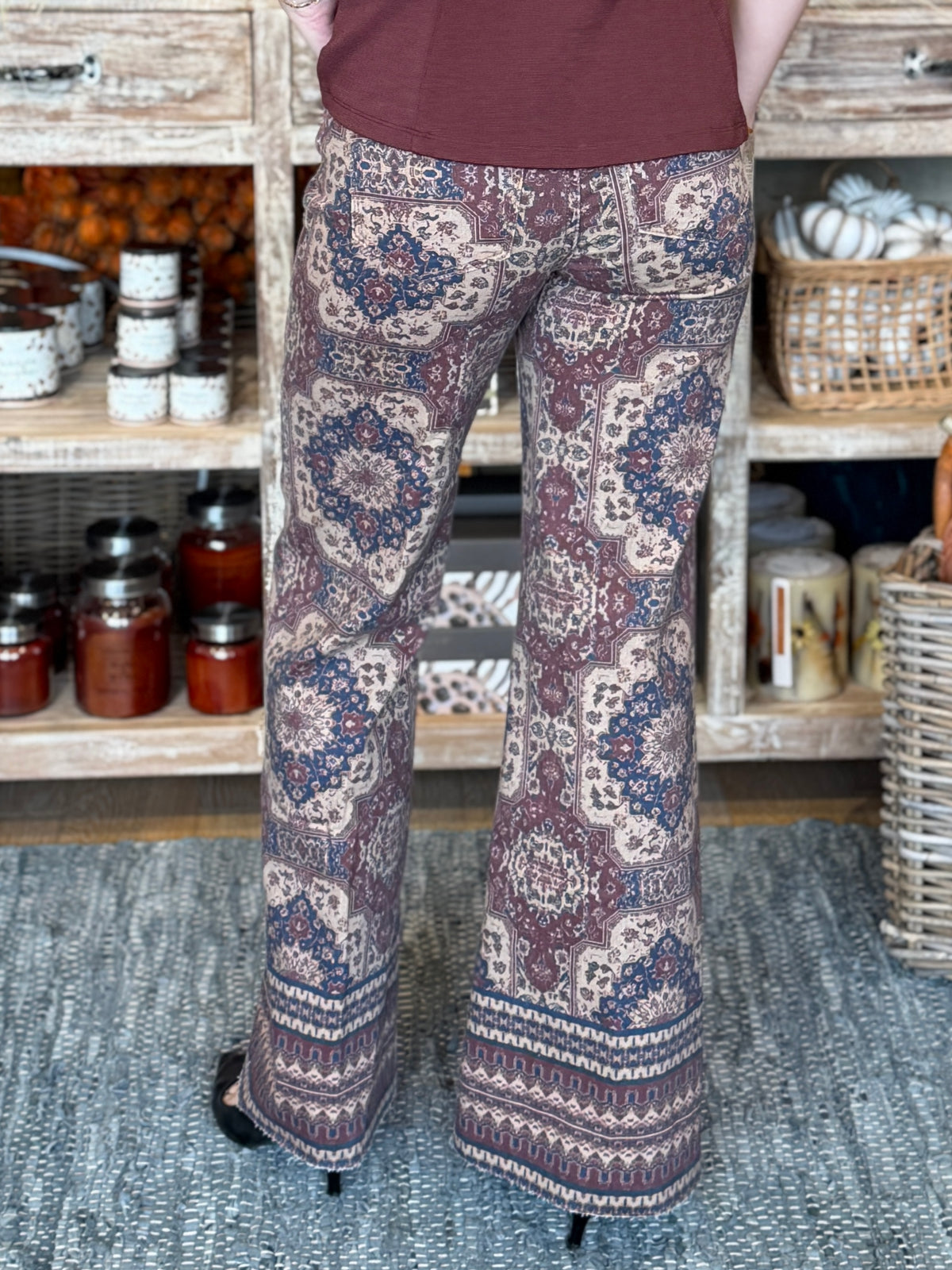 A person shown from the back wearing patterned wide-leg pants with a rust brown top, standing on a textured rug in front of rustic wooden shelves filled with jars and baskets.