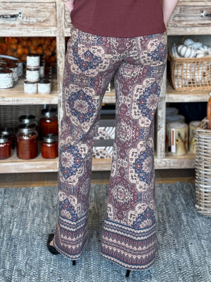 A person shown from the back wearing patterned wide-leg pants with a rust brown top, standing on a textured rug in front of rustic wooden shelves filled with jars and baskets.