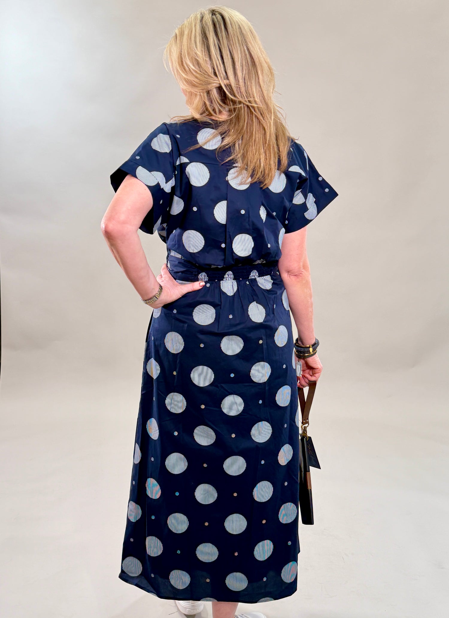 Rear view of a woman in a navy polka dot 100% cotton midi dress with a removable belt and white sneakers