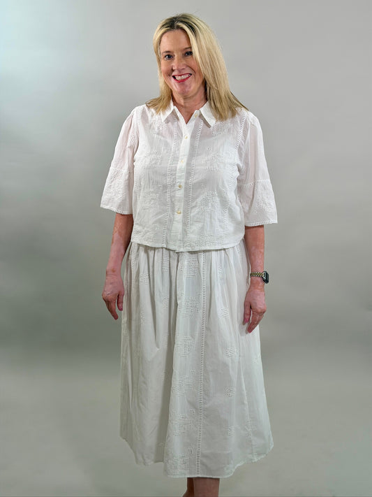 A smiling blonde woman wearing a white geometric eyelet cropped button-up shirt with a matching maxi skirt.