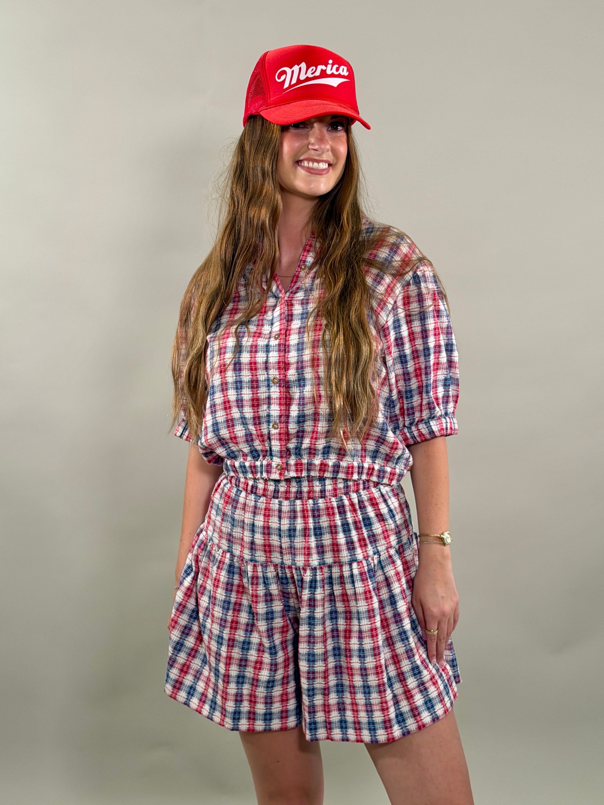 Woman smiling and wearing a red, navy, and white plaid short-sleeve cropped button-down shirt with a matching skort and a red baseball cap.