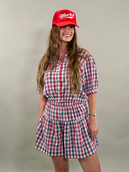 Woman smiling and wearing a red, navy, and white plaid short-sleeve cropped button-down shirt with a matching skort and a red baseball cap.