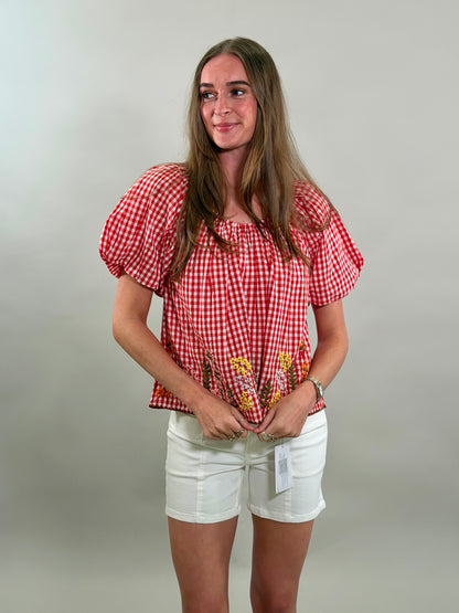 Woman looking to the side wearing a red checkered blouse with puff sleeves and white shorts against a plain background