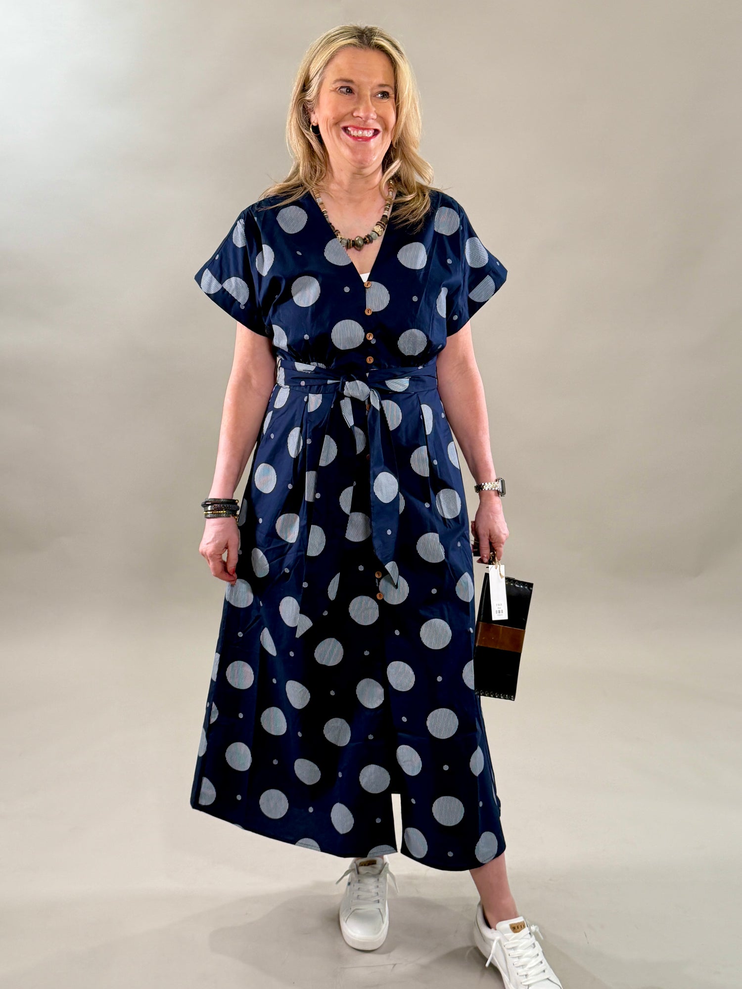 Woman smiling and looking to the side in a navy polka dot 100% cotton midi dress with a removable belt and white sneakers