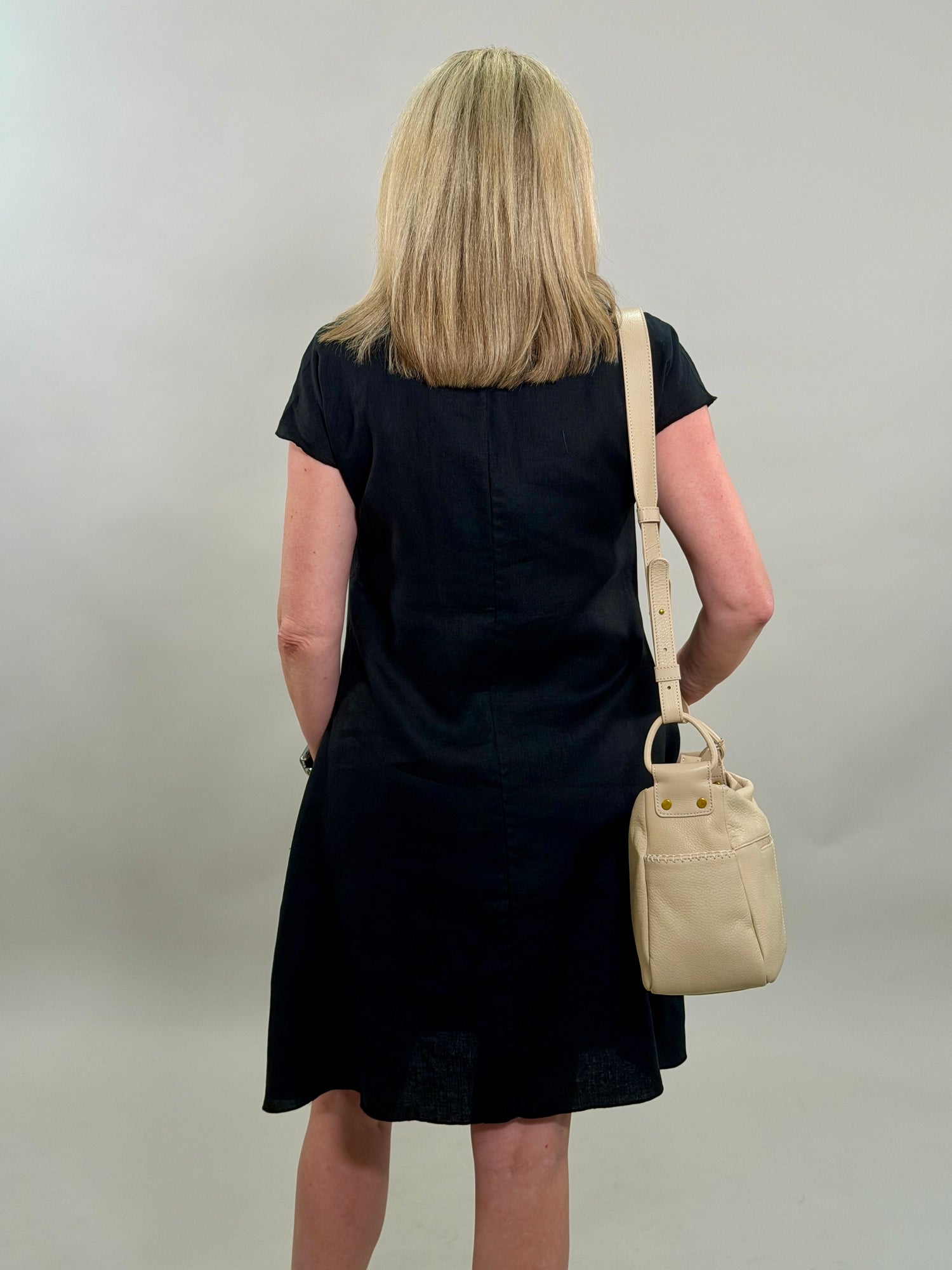Rear view of a blonde woman wearing a classic black 100% linen cowl-neck midi dress with a tan crossbody bag and hand in her pocket.