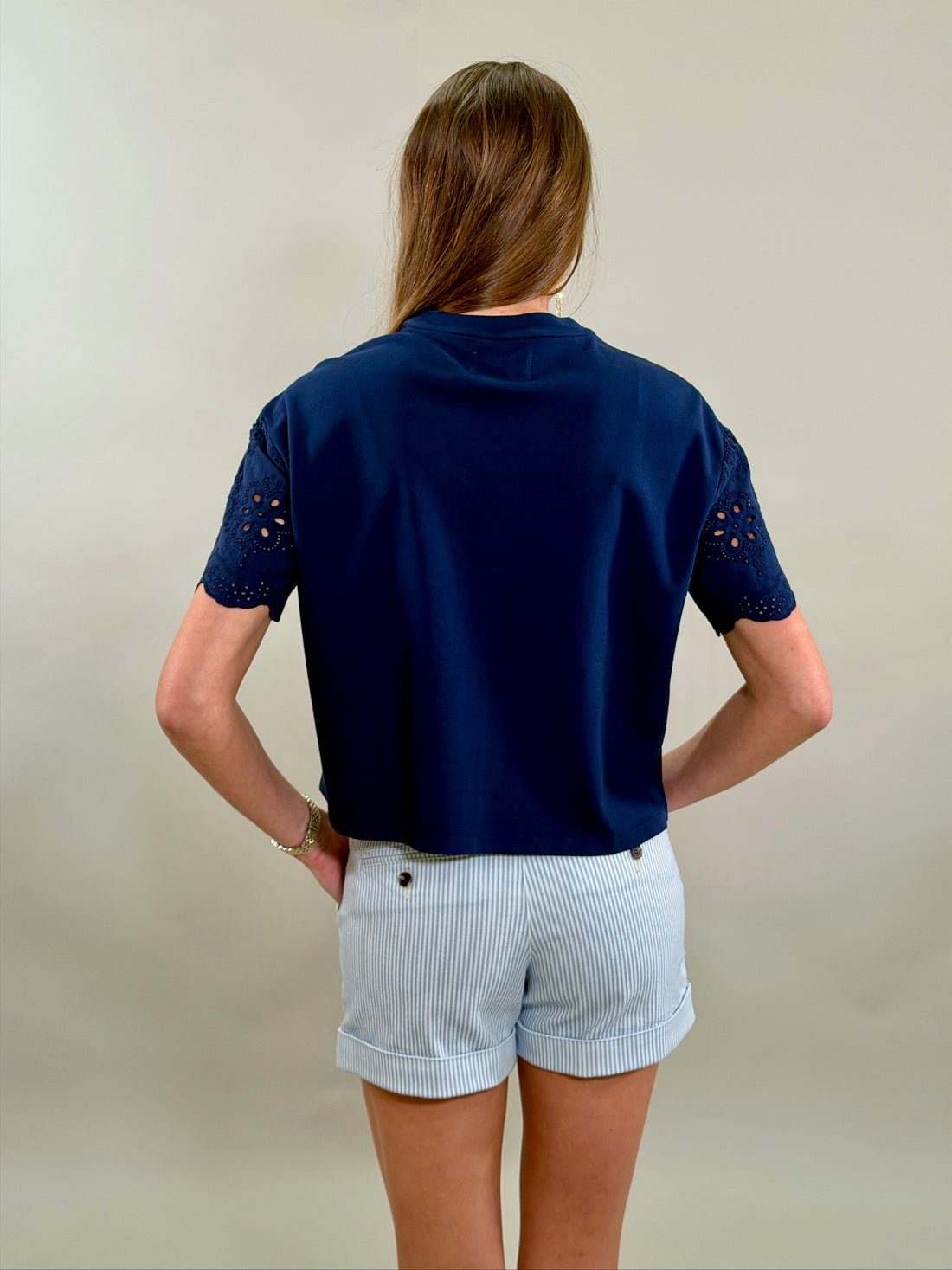 Rear view of a woman wearing a navy blue top with eyelet sleeves and light blue pinstripe shorts on a plain background