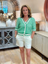 Woman standing in a boutique wearing a green and cream striped open-knit short-sleeve cardigan sweater with white shorts.