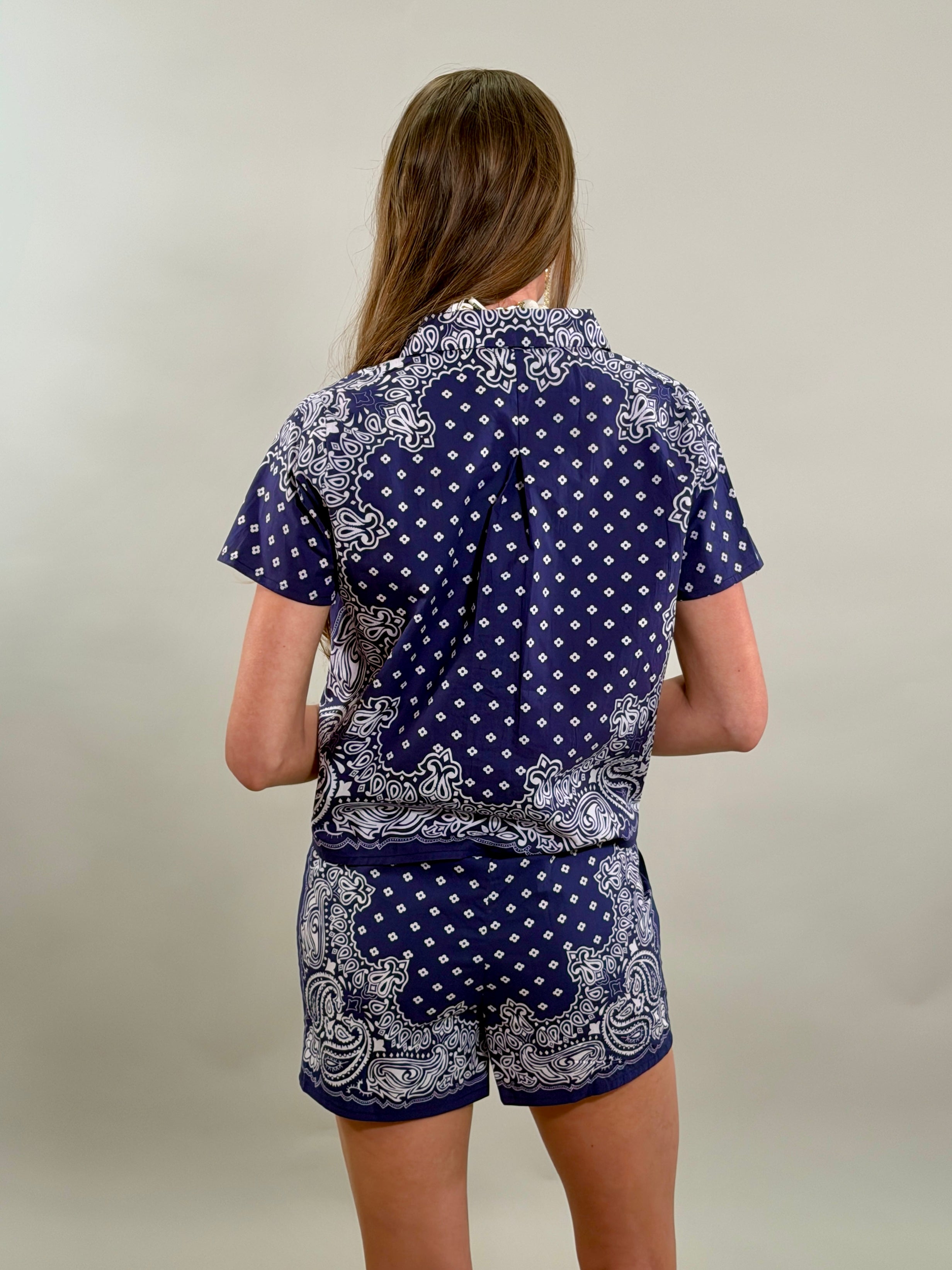 Rear view of a woman with long brown hair, showing the back of a navy blue and white paisley bandana-print short-sleeve shirt.