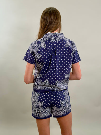 Rear view of a woman with long brown hair, showing the back of a navy blue and white paisley bandana-print short-sleeve shirt.
