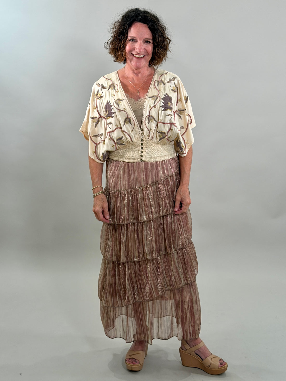 A smiling woman wearing a cream-colored bohemian blouse with purple and gold floral embroidery, a smocked waist, and a tan lace bralette underneath.