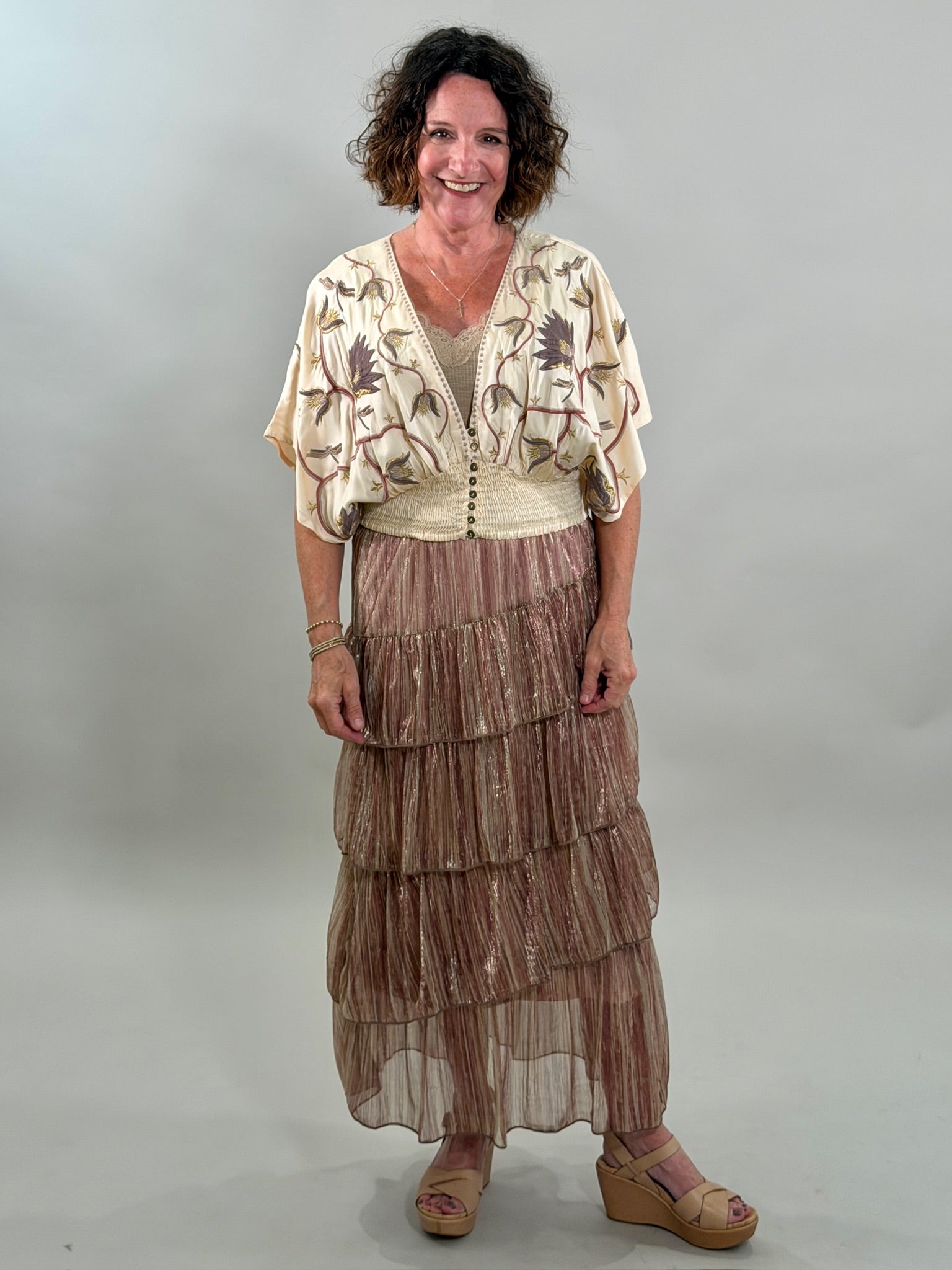 A smiling woman wearing a cream-colored bohemian blouse with purple and gold floral embroidery, a smocked waist, and a tan lace bralette underneath.