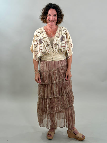A smiling woman wearing a cream-colored bohemian blouse with purple and gold floral embroidery, a smocked waist, and a tan lace bralette underneath.