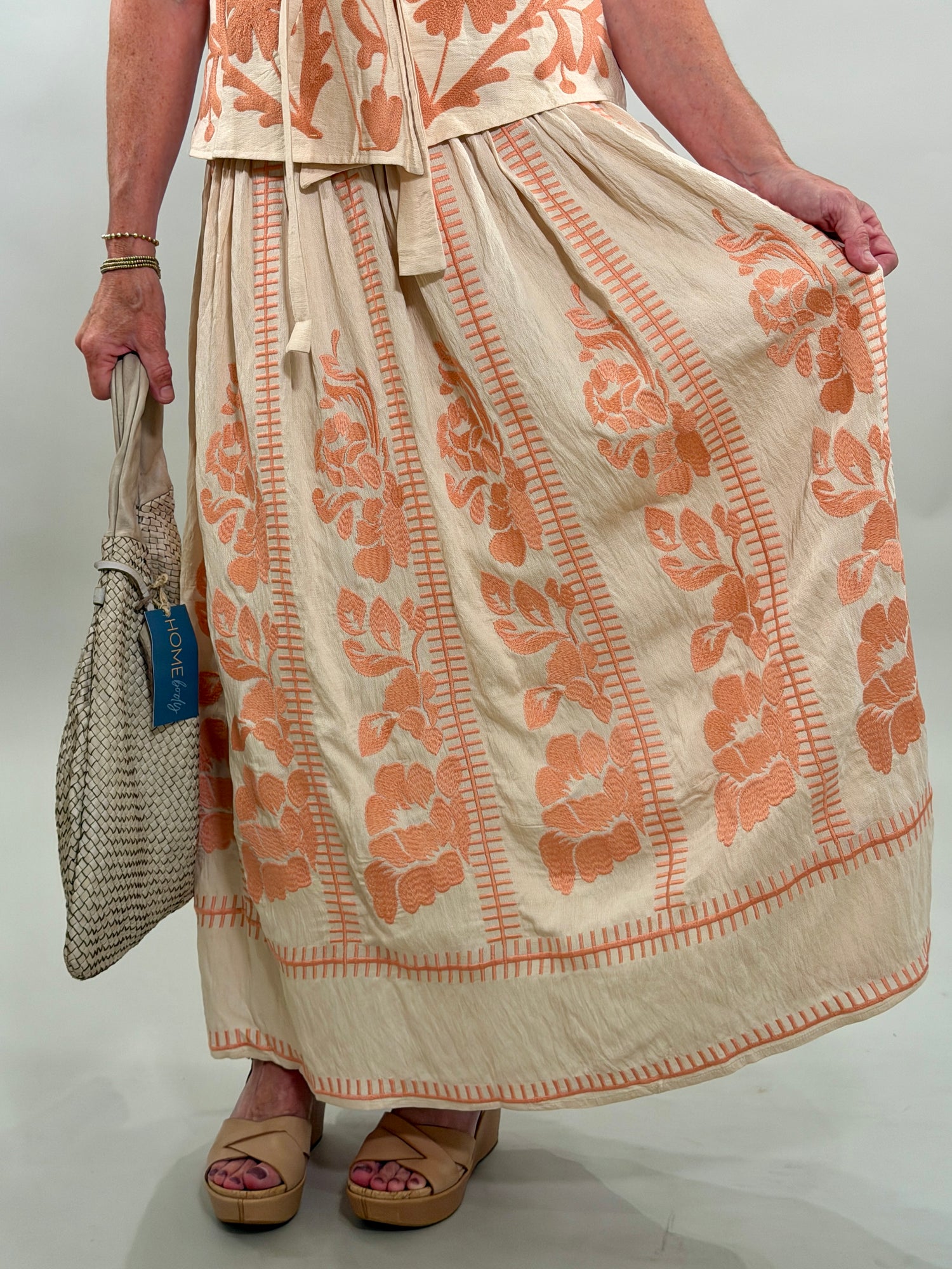 Close up of a woman wearing a long beige skirt with coral floral patterns, holding a woven bag.