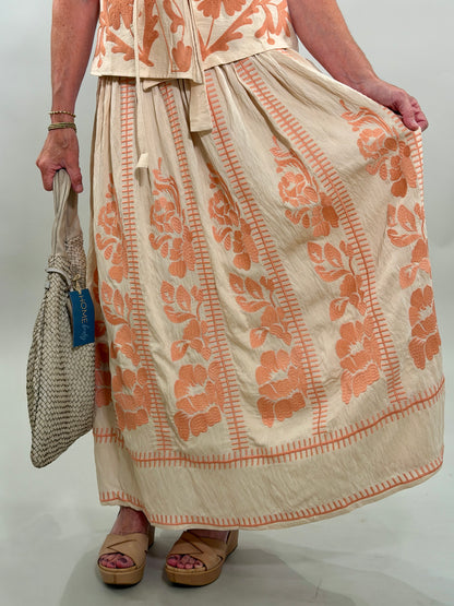 Close up of a woman wearing a long beige skirt with coral floral patterns, holding a woven bag.