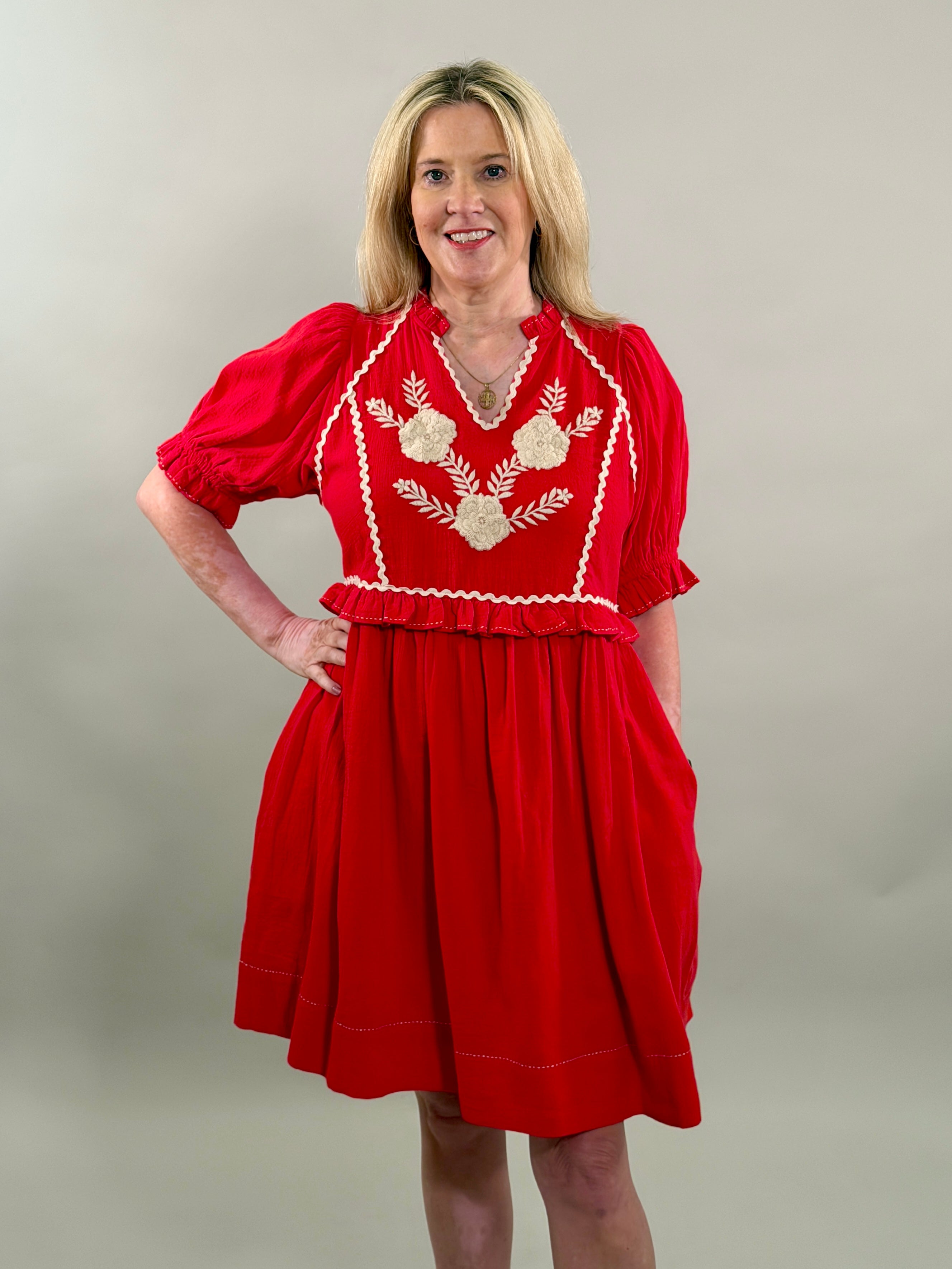 A smiling blonde woman with her hand on her hip wearing a vibrant red 100% cotton dress with cream-colored floral embroidery and puff sleeves.