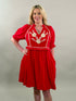 A smiling blonde woman with her hand on her hip wearing a vibrant red 100% cotton dress with cream-colored floral embroidery and puff sleeves.