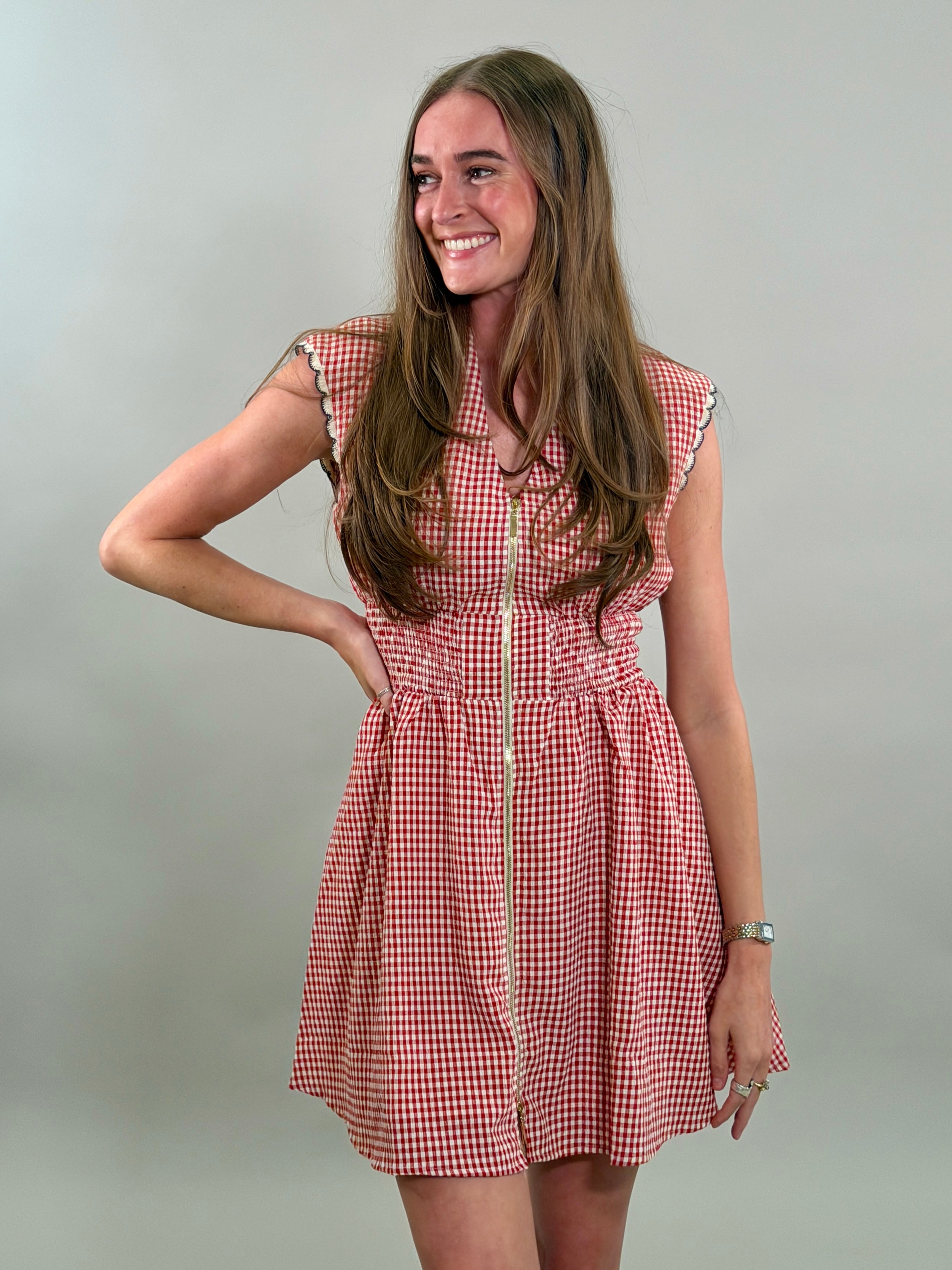 A smiling woman with long brown hair wearing a red and white gingham sleeveless dress with a gold front zipper and a smocked waistband.