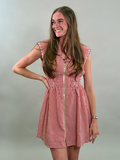 A smiling woman with long brown hair wearing a red and white gingham sleeveless dress with a gold front zipper and a smocked waistband.