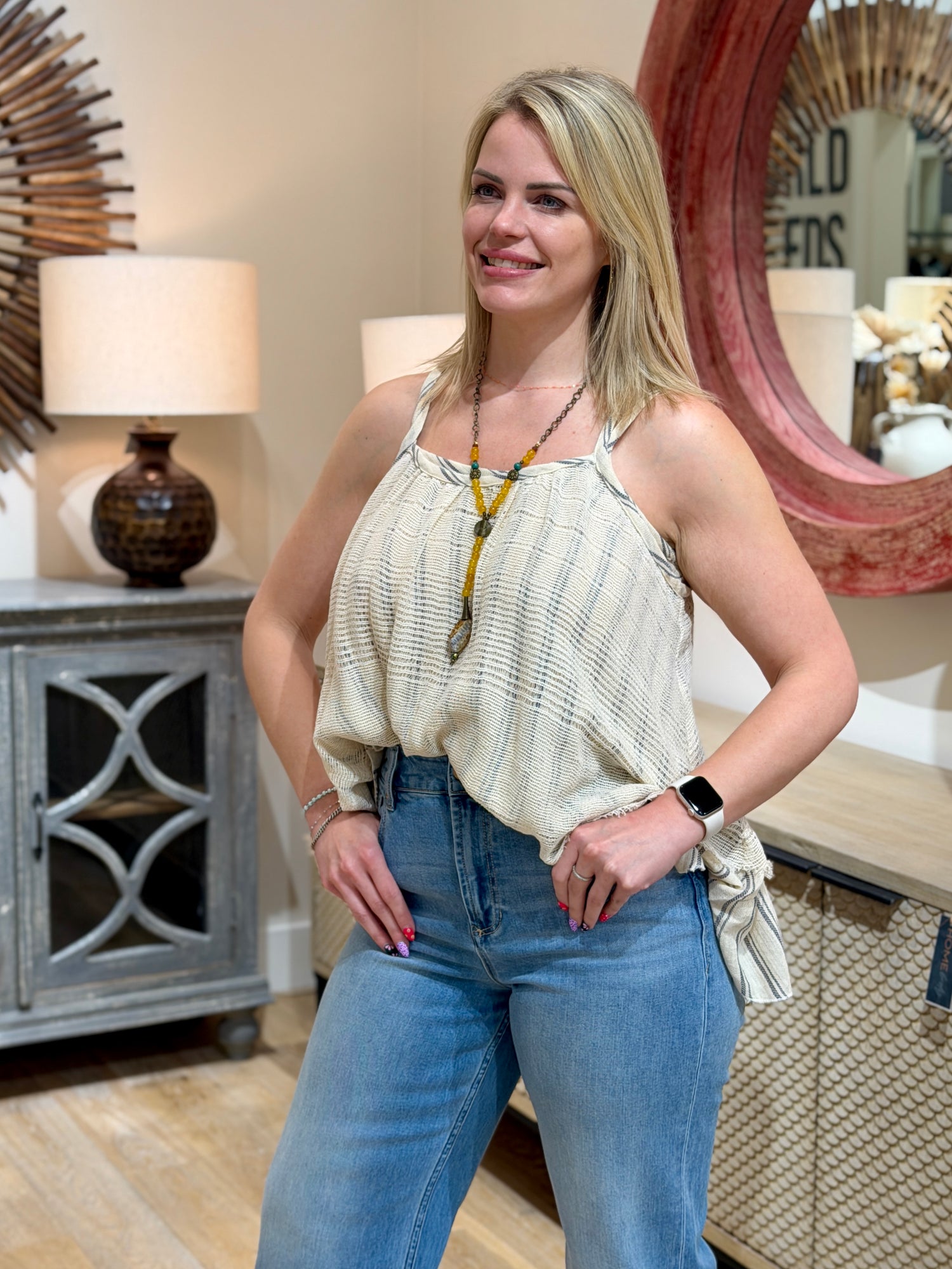 Close up view of a woman wearing a cream and grey textured striped cami tank top tucked into blue jeans.