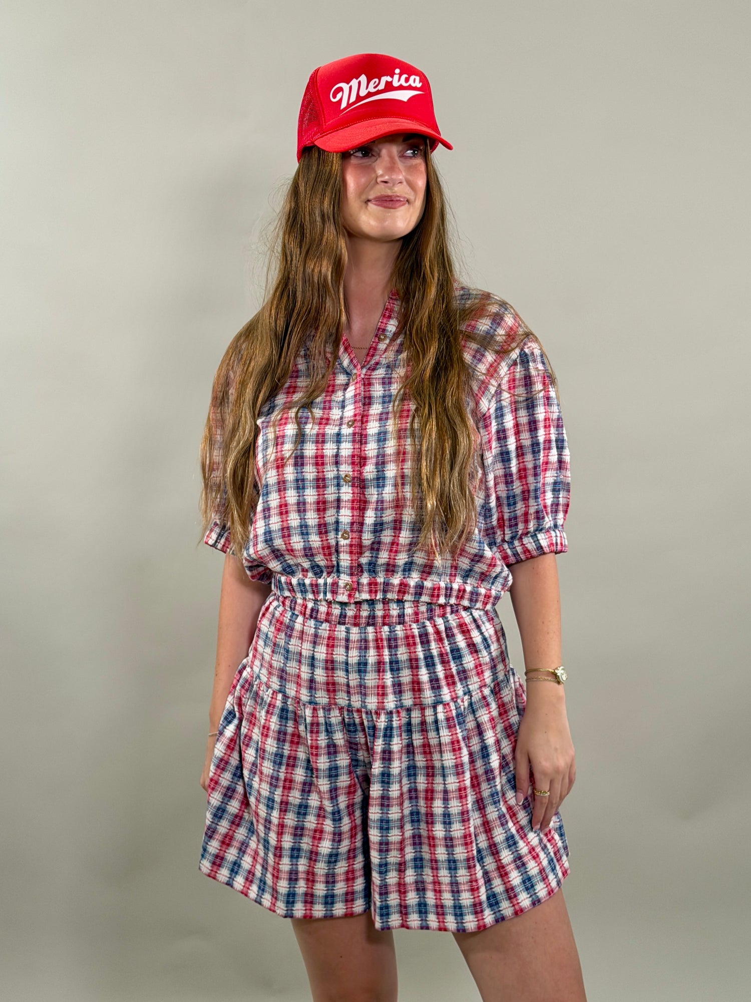 Woman smiling and looking to the side, wearing a red, navy, and white plaid short-sleeve cropped button-down shirt with a matching skort and a red baseball cap.