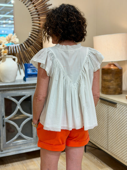 Rear view of a woman with curly hair and glasses smiling while wearing a white cotton flutter sleeve top with orange floral embroidery and matching orange shorts.