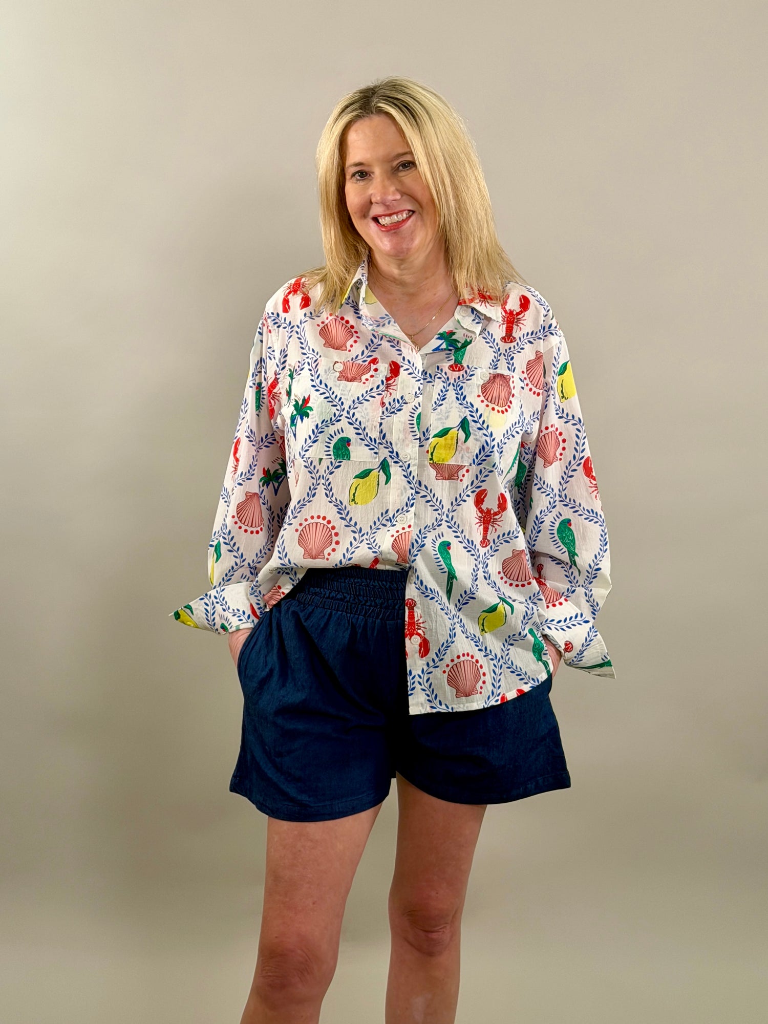 Smiling woman wearing an oversized white button-down shirt with a vibrant blue, red, and yellow coastal print paired with navy shorts.