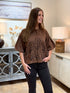 A woman modeling a brown short-sleeve top with black floral embroidery, paired with black pants, standing in a brightly lit home interior.