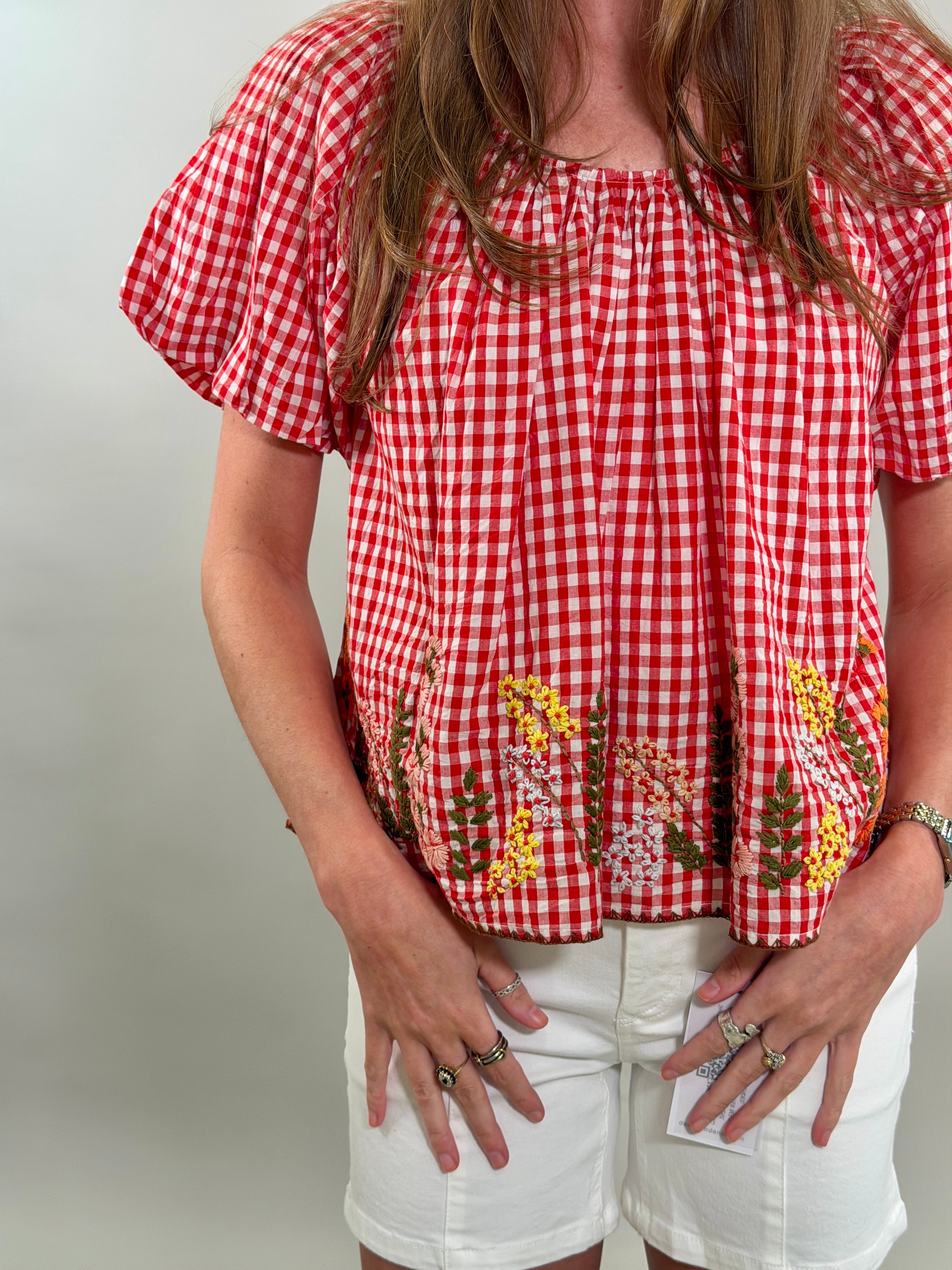 Close up of a woman wearing a red and white checkered blouse with floral embroidery on a plain background