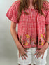 Close up of a woman wearing a red and white checkered blouse with floral embroidery on a plain background