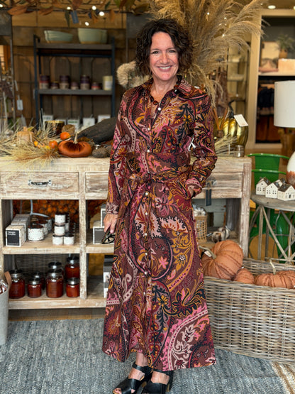 A woman with curly dark hair stands in a rustic home decor store wearing a long-sleeved, button-front maxi dress with a rich paisley pattern in deep red, brown, orange, and black. She is standing in front of a wooden display cabinet filled with home decor items, pumpkins, and baskets.