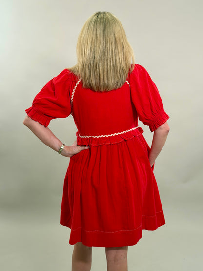 Rear view of a blonde woman with her hand on her hip wearing a vibrant red 100% cotton dress with cream-colored ricrac embroidery and puff sleeves.