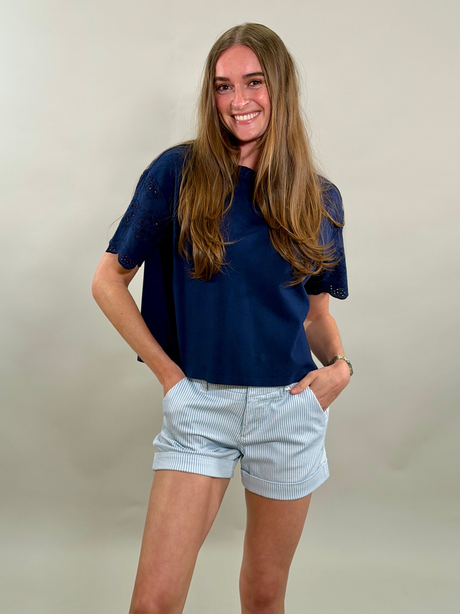A smiling woman wearing a navy blue top and light blue pinstripe shorts against a plain background