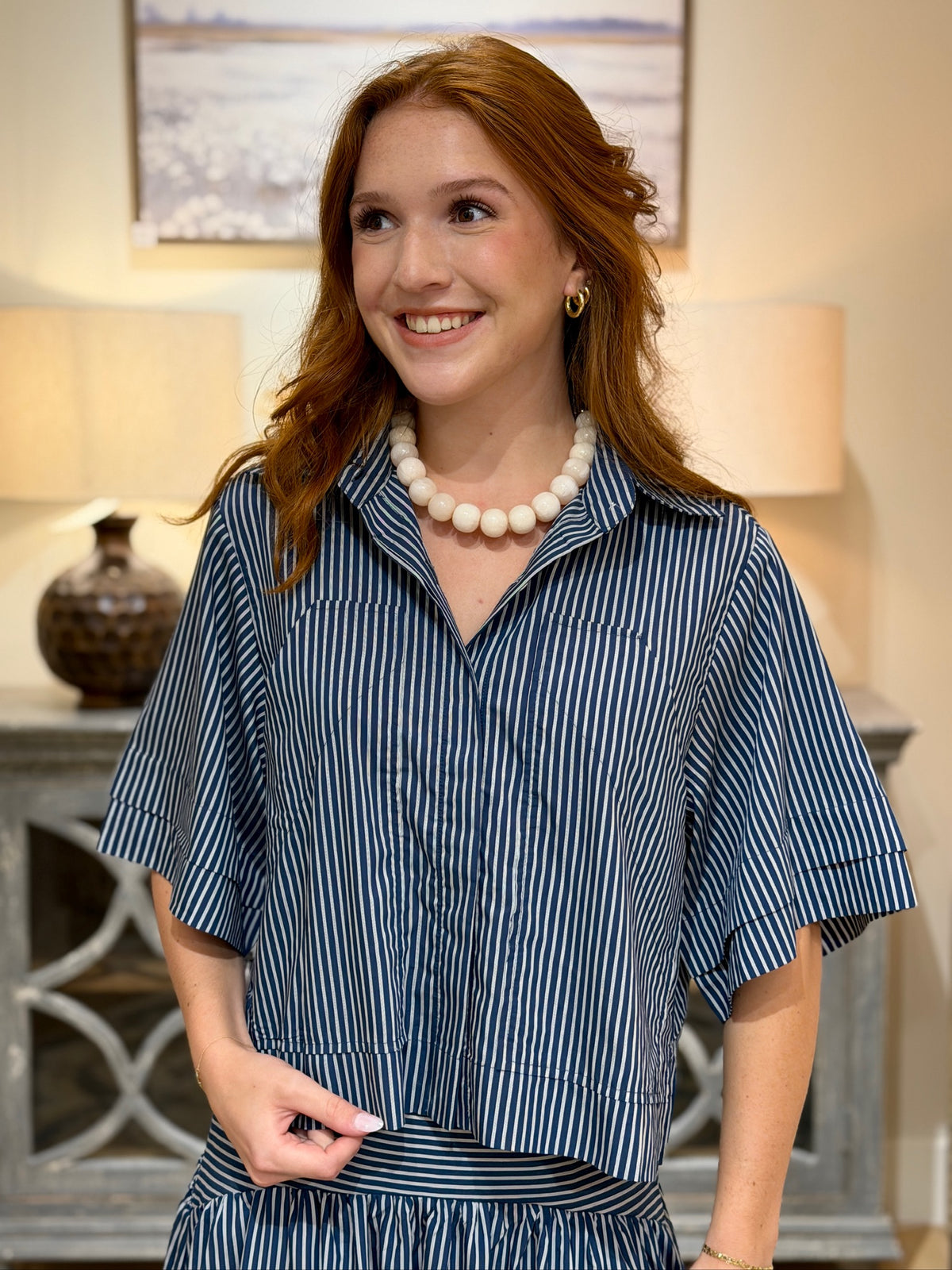 A smiling woman with red hair wearing a blue and white vertically striped short-sleeve collared shirt and matching skirt, styled with a white beaded necklace in a warmly lit indoor setting.