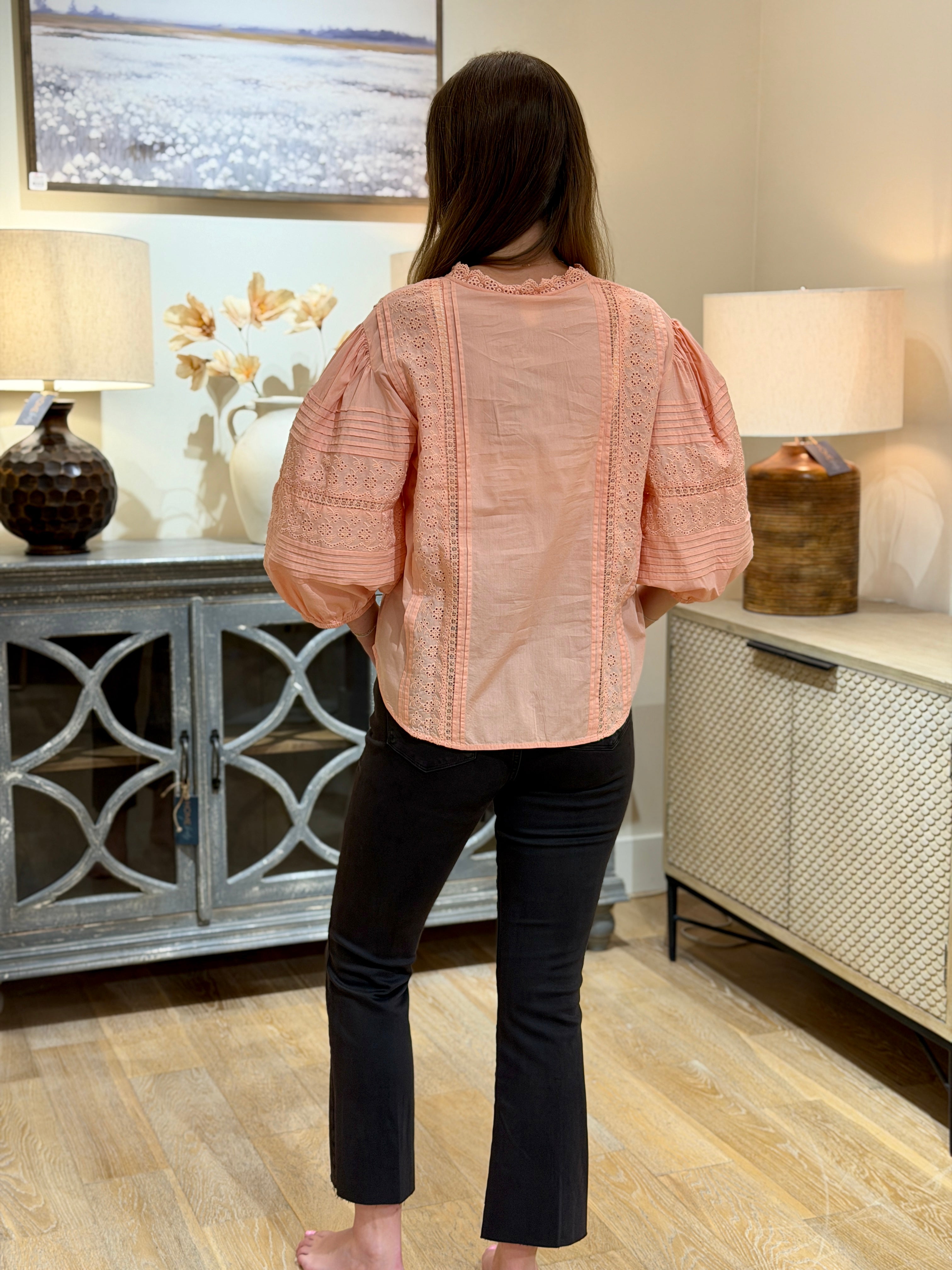 A woman viewed from behind wearing a peach-pink short-sleeve blouse with lace embroidery and puff sleeves, paired with black pants, standing in a styled room with wooden floors and sideboards.