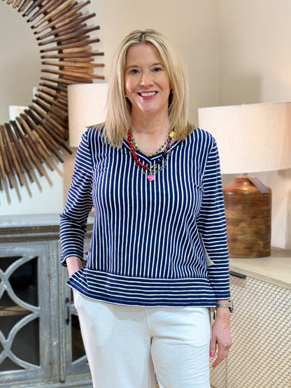 Close up of a blonde woman smiling while wearing a navy and white striped cotton-linen V-neck pullover top with white trousers and a colorful necklace.