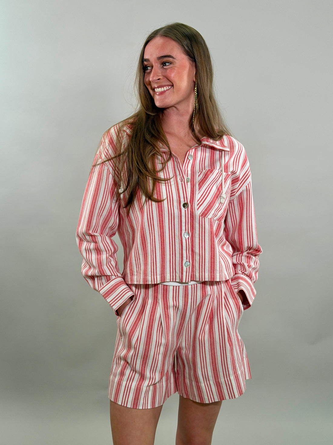 A smiling woman with long brown hair stands with her hands in her pockets, wearing a relaxed matching set featuring a red and white candy-striped cropped long-sleeve button-down shirt and matching pleated shorts.