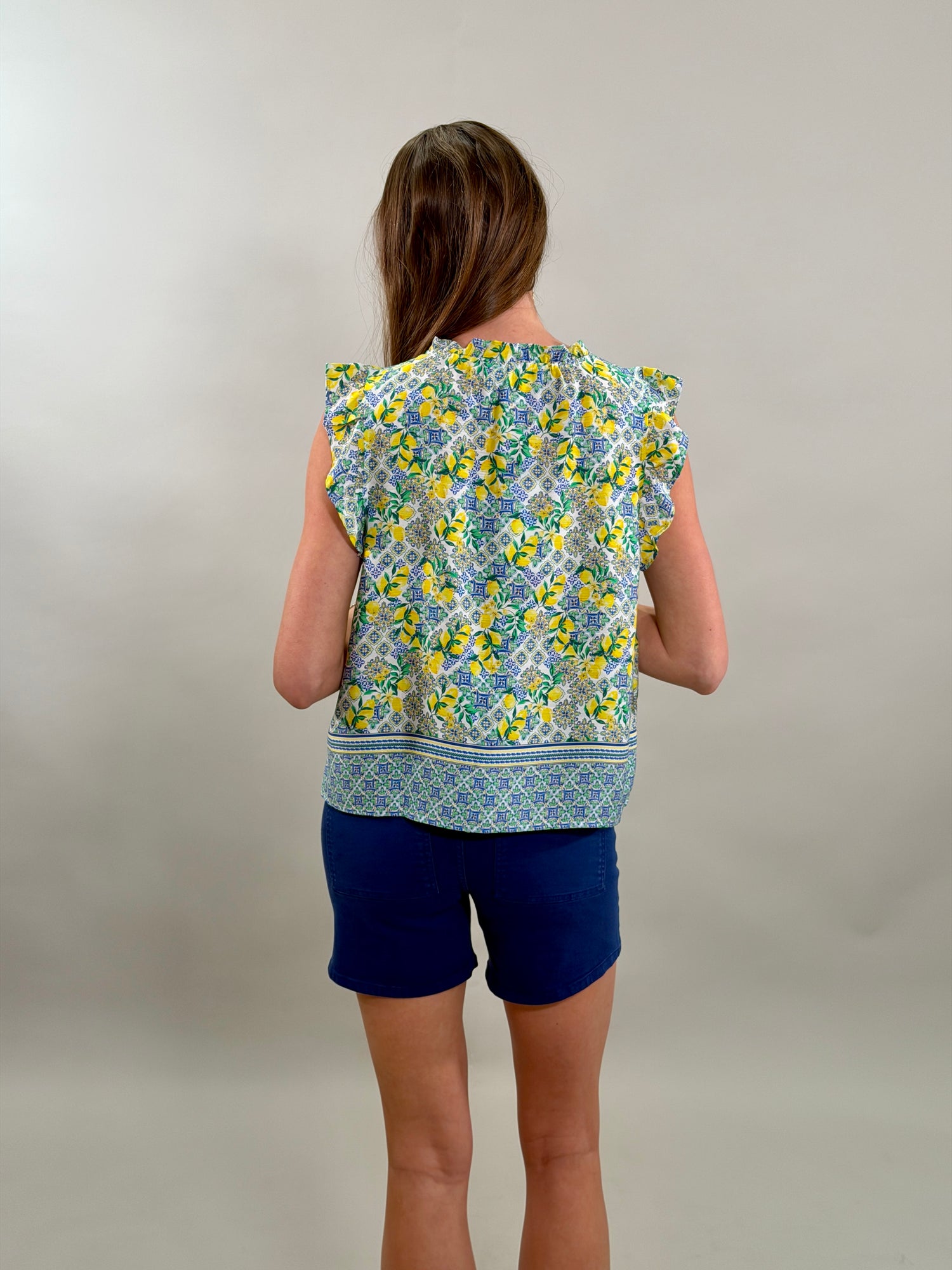 Rear view of a woman wearing a sleeveless white blouse with a blue tile and yellow lemon print paired with royal blue shorts.