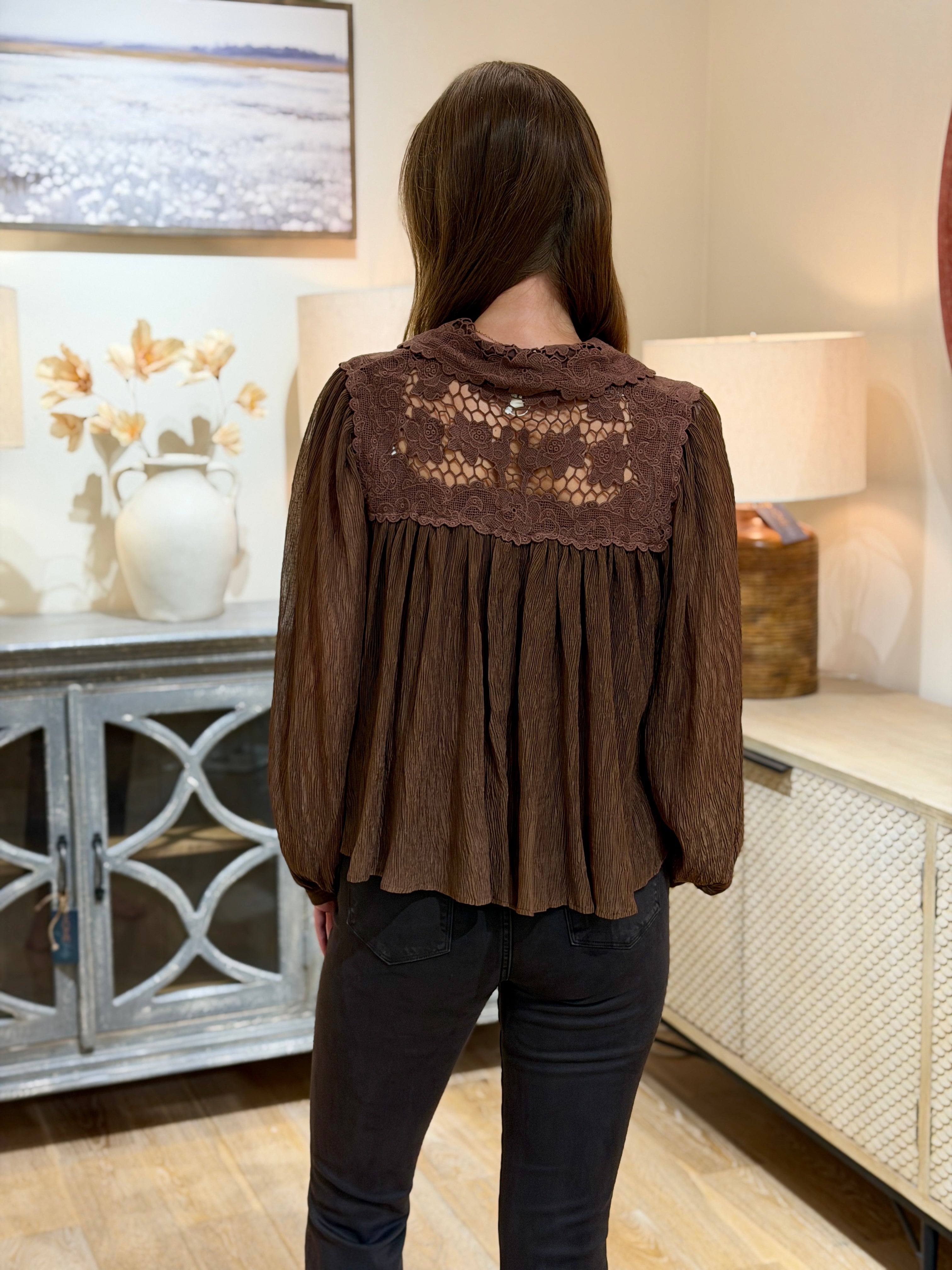 A woman viewed from behind wearing a brown long-sleeve blouse with a lace yoke and dark pants, standing in a home interior setting.