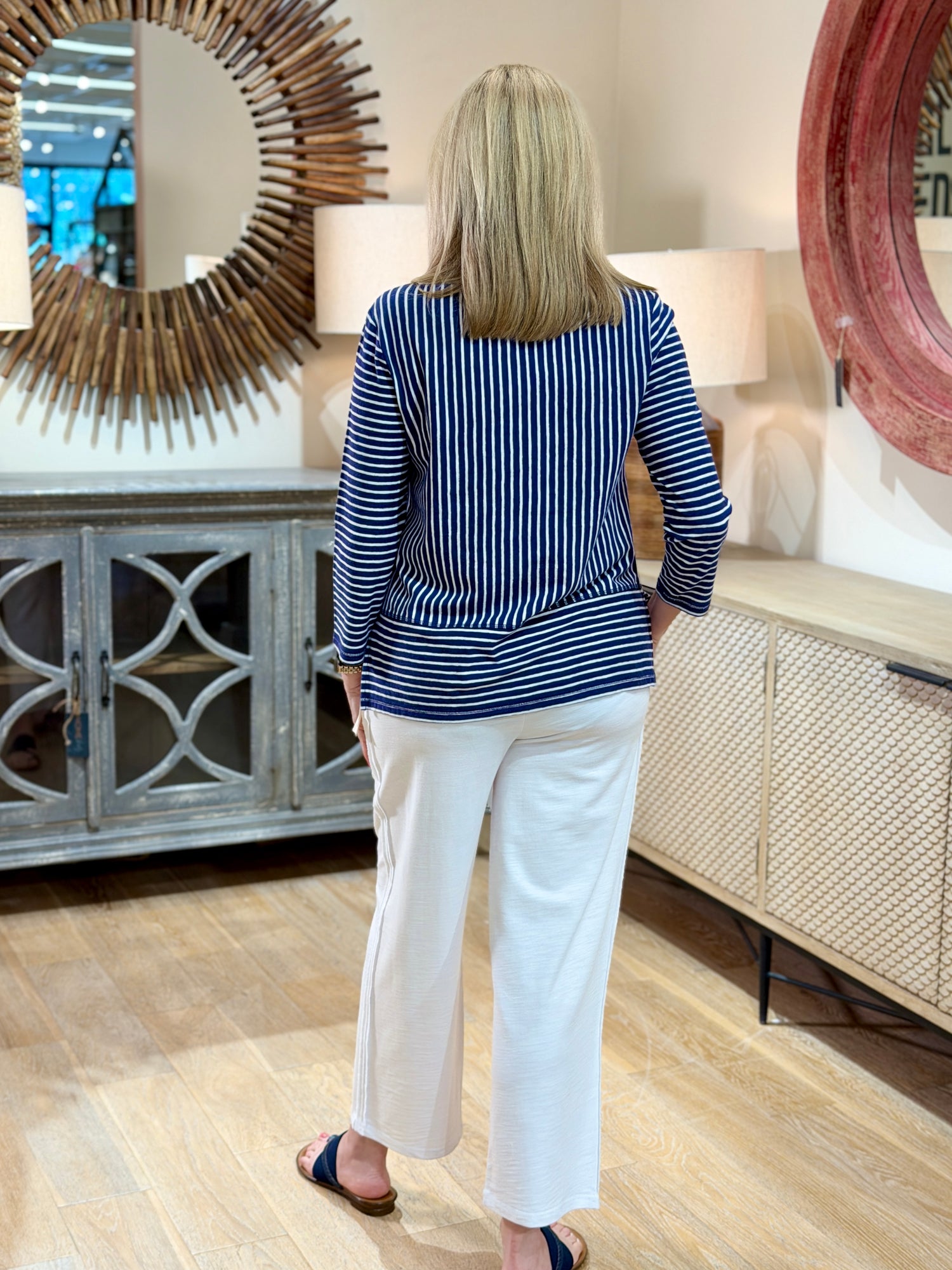Rear full body view of a blonde woman wearing a navy and white striped cotton-linen V-neck pullover top with white trousers.