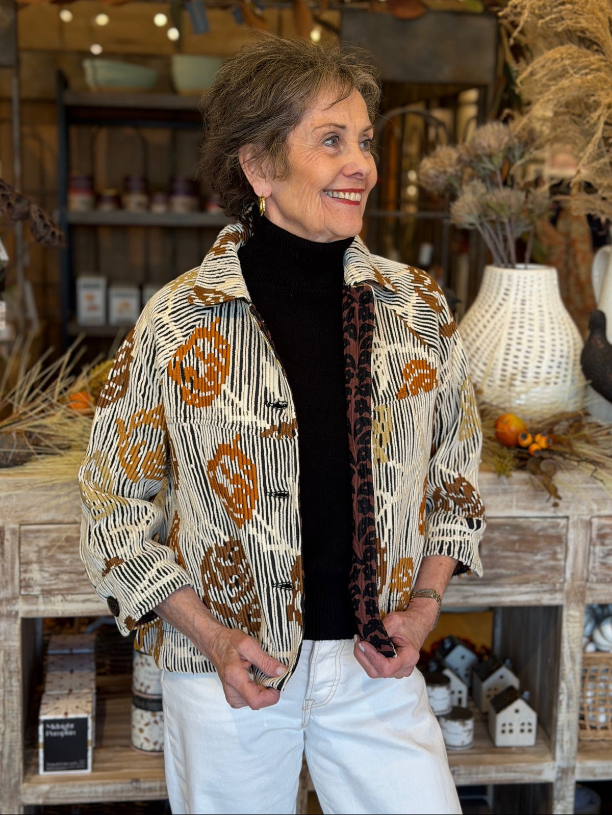 A smiling woman wearing a patterned button-up shirt jacket over a black turtleneck and white pants, standing in a rustic home decor store.