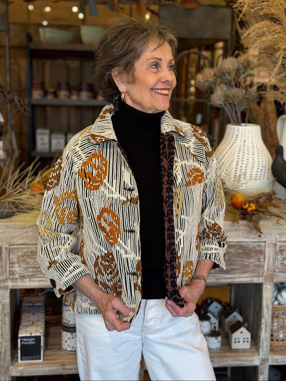 A smiling woman wearing a patterned button-up shirt jacket over a black turtleneck and white pants, standing in a rustic home decor store.