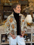 A smiling woman wearing a patterned button-up shirt jacket over a black turtleneck and white pants, standing in a rustic home decor store.