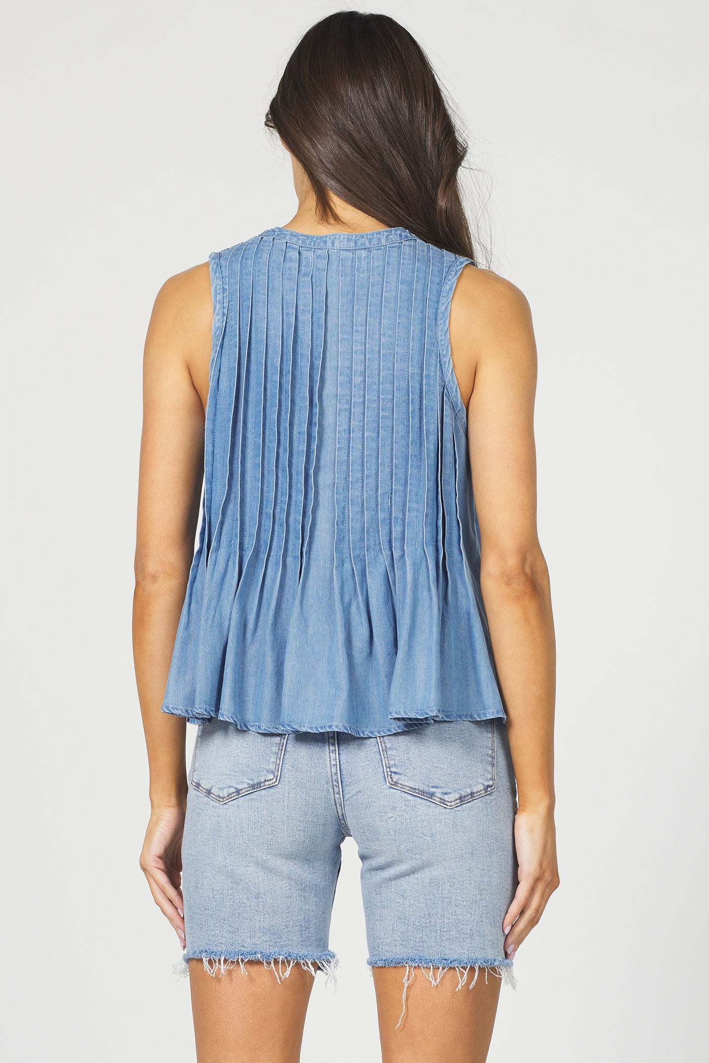 A rear view of a woman wearing the sleeveless pleated Tencel tank top in denim blue with light-wash denim shorts